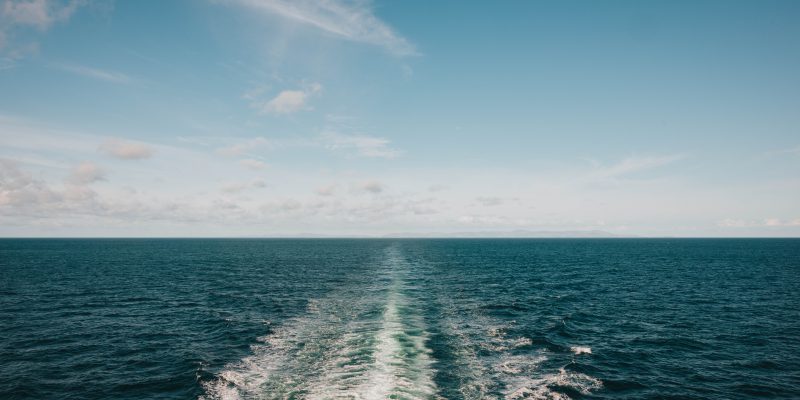 The wake of a vessel seen as white lines in a relatively calm blue sea, looking to a distant cloudy horizon