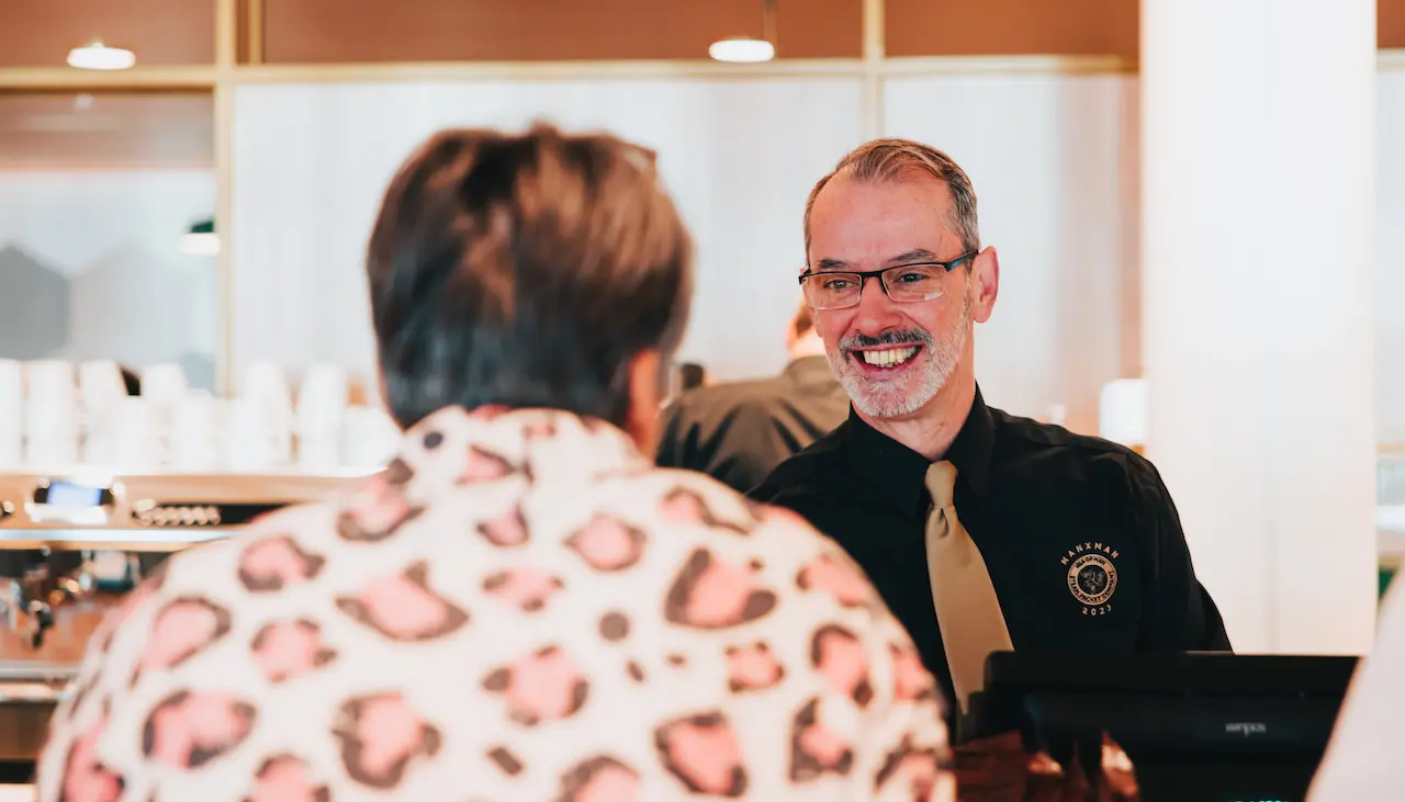 A friendly barista with a beard, wearing black and gold Manxman uniform serves a customer at the Cornaa Cafe on deck 7 of Manxman