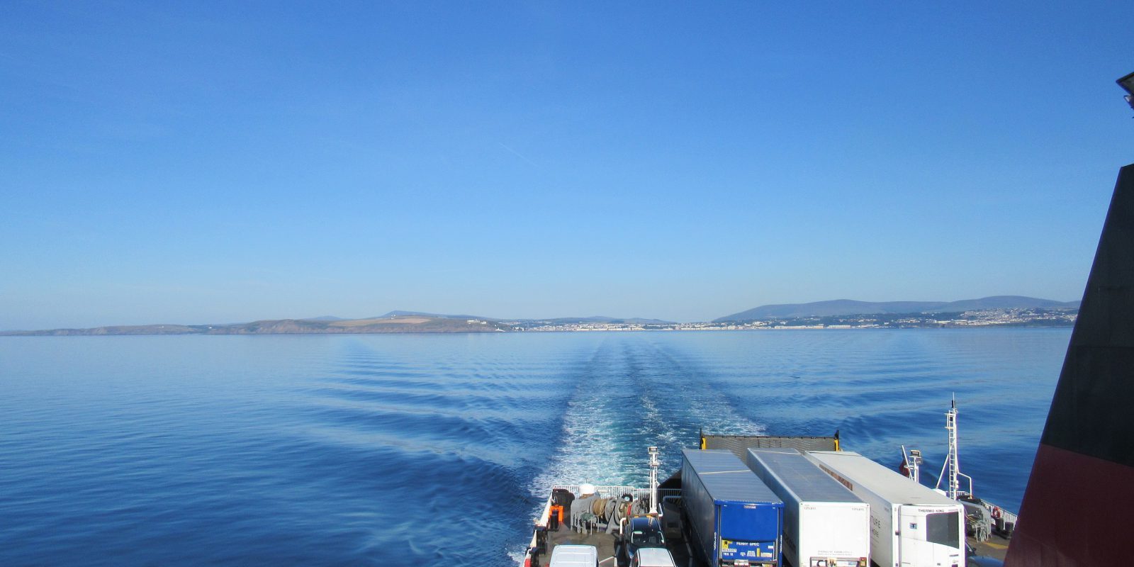 View from back of ferry vessel with lorry's on the ferry in the foreground and the sea and coastline in the background.