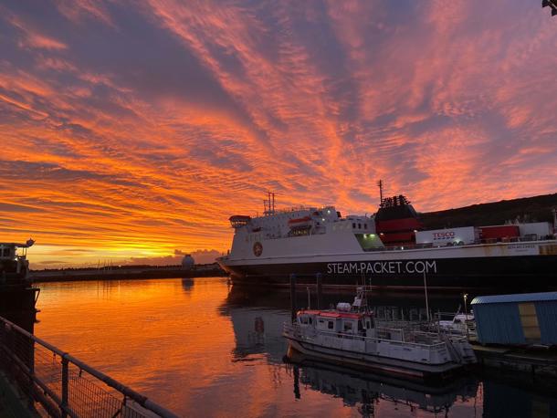 A photo of Steam Packet Company vessel Ben-my-Chree in Douglas Harbour with a beautiful orange sunrise reflection which reflects onto the calm waters with a small fishing boat in the foreground