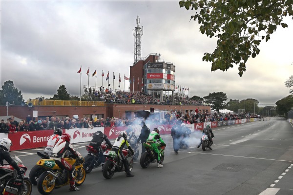 Start line of a motorcyle race with motorcyclists lined up, and the crowd in the stands in the background