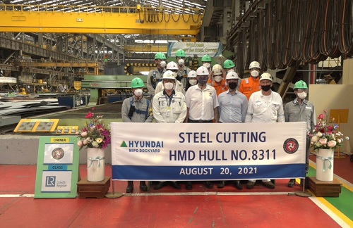 Steel cutting staff in ship construction warehouse behind a banner that says " Hyundai MIPO Dockyard, Steel Cutting HMD Hull No.8311, August 20, 2021”