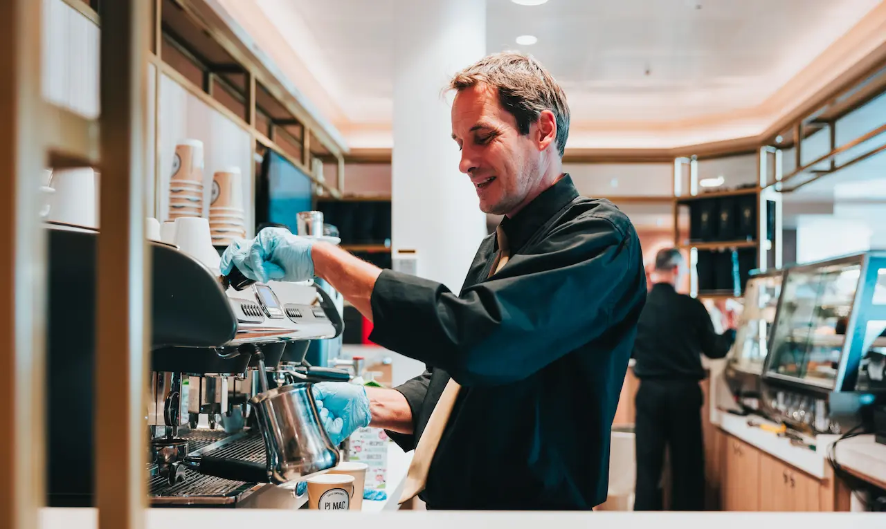 A barista, dressed in a black uniform and wearing disposable food hygiene gloves, froths milk in a stainless steel jug while his colleague serves a customer in the background.